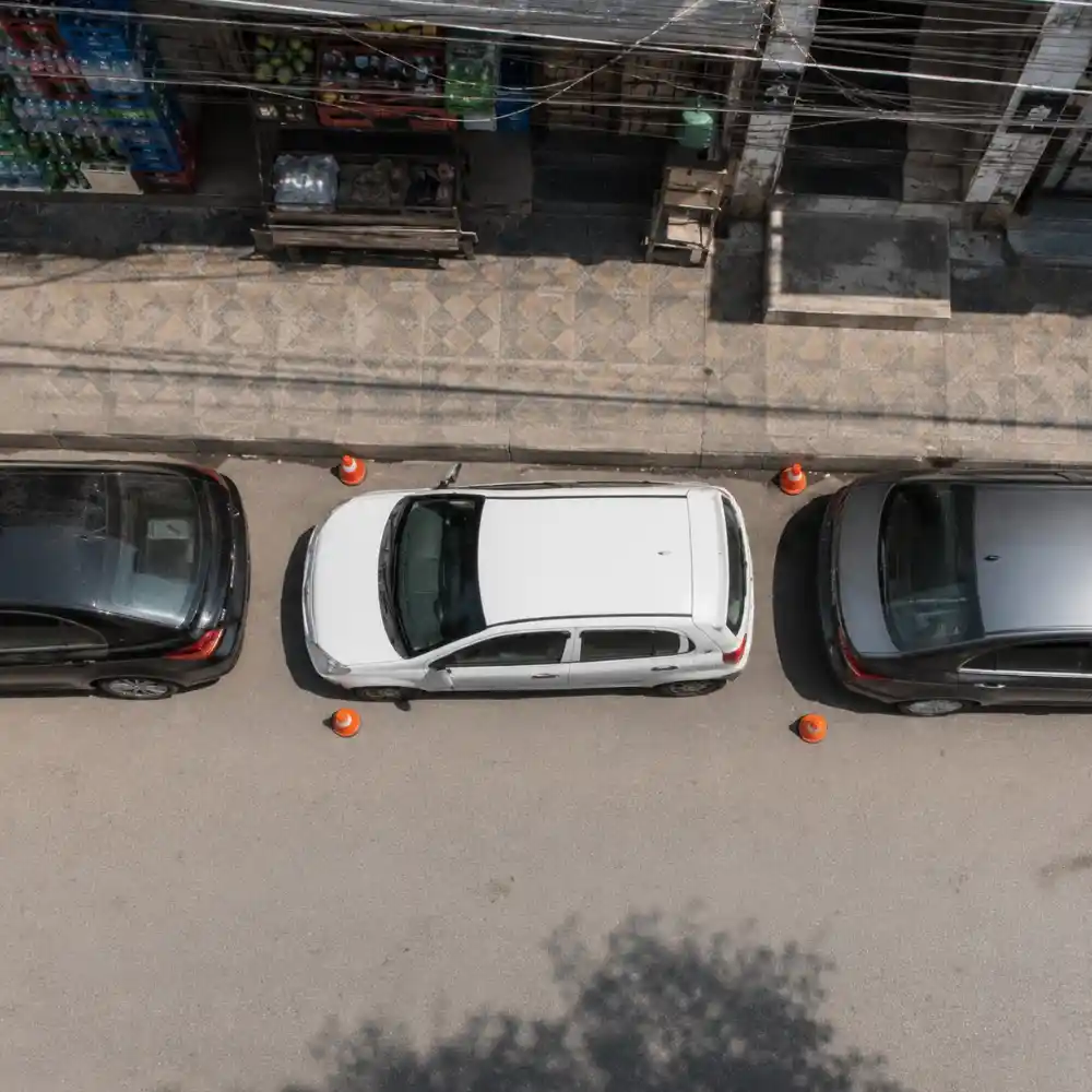 Aerial view of a white car doing parallel parking between two cars with orange traffic cones during advanced driving training at The Dreamial Driving School Lahore.